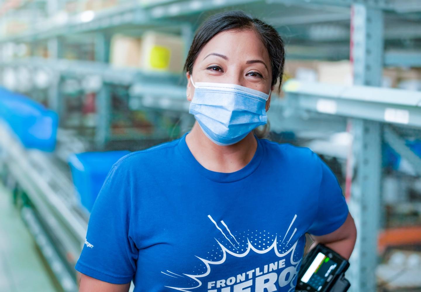 A Cencora team member smiling while wearing a blue surgical mask in a warehouse. They are wearing a blue t-shirt that reads “Frontline Hero.”